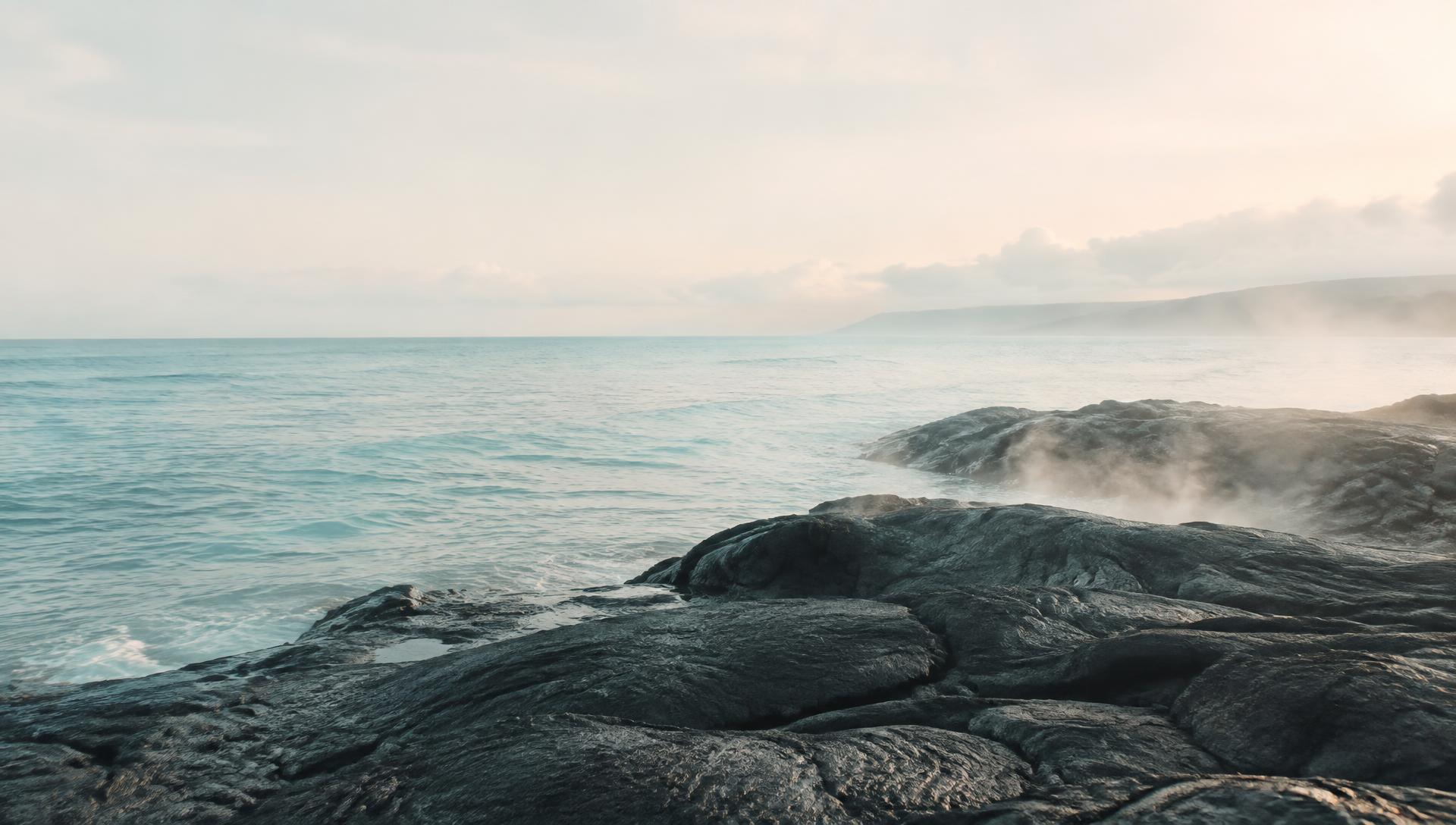 Gentle steam rising from black lava rock where it meets a calm tropical ocean